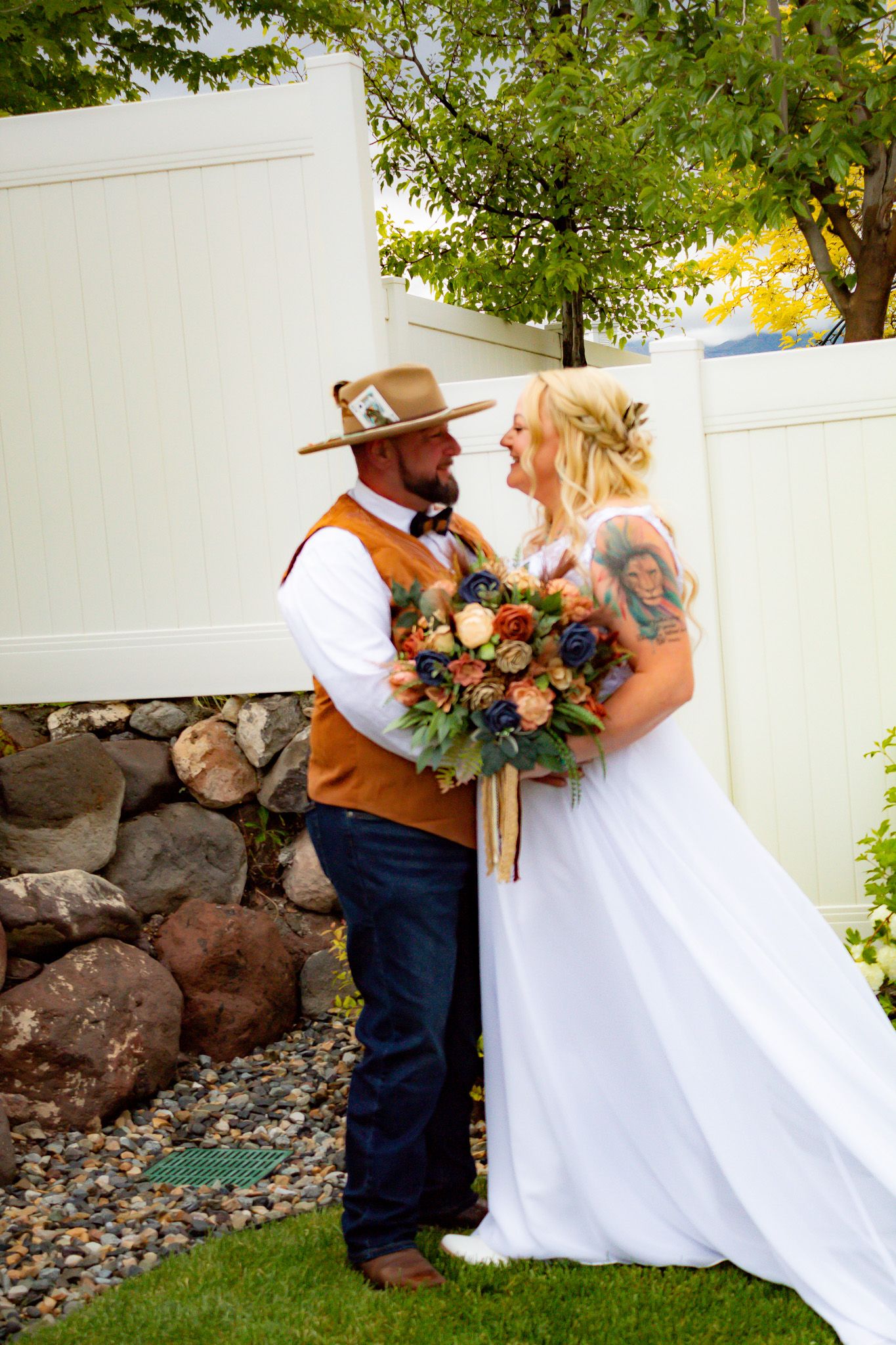 Newlyweds embrace in garden setting with white fence and autumn foliage.