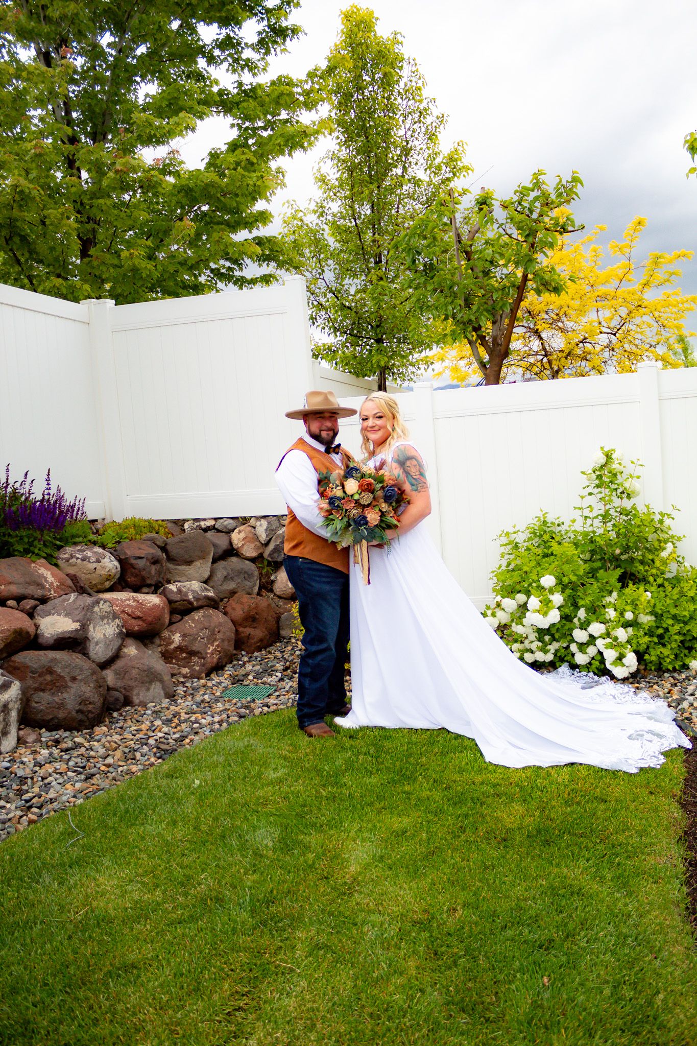 Wedding couple poses in landscaped garden with stone wall and white fence backdrop.