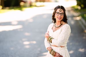 Bride in lace dress and glasses poses outdoors in dappled sunlight.