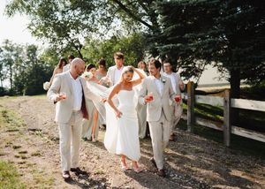 Wedding party in white and cream attire walking along a rustic fence path.