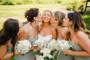 Bridesmaids in sage green dresses sharing joyful moments with bouquets outdoors.