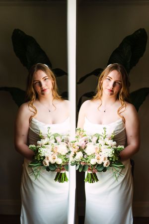 Mirror reflection of bride holding white and pink floral bouquet against dark background.