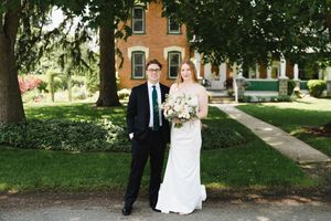 Wedding couple poses for portrait on tree-lined path in front of brick building.