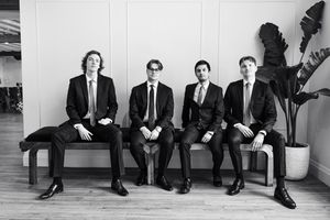 Groomsmen in black suits sit casually against a white wall in a modern black and white photo.