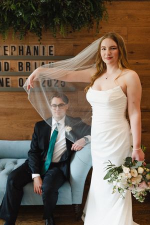 Bride shows off veil while groom sits on blue couch in rustic wooden venue.