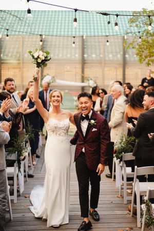 Newlyweds celebrate while walking down the aisle under string lights at an outdoor wedding ceremony.
