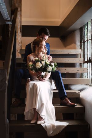 Couple sits together on rustic wooden stairs in romantic window light.