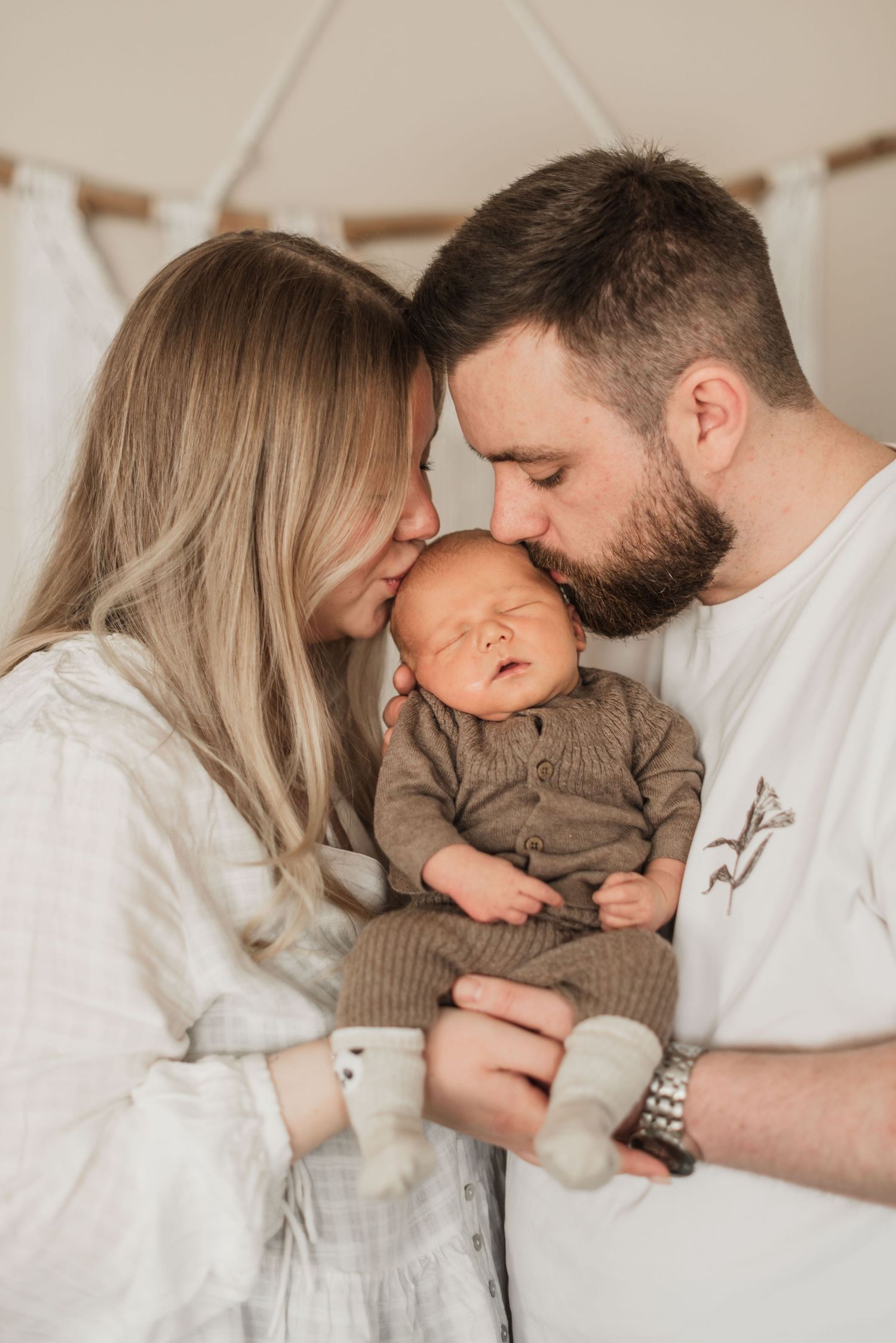 A couple tenderly kisses their sleeping newborn baby, dressed in olive green, in a warm, softly lit nursery setting.