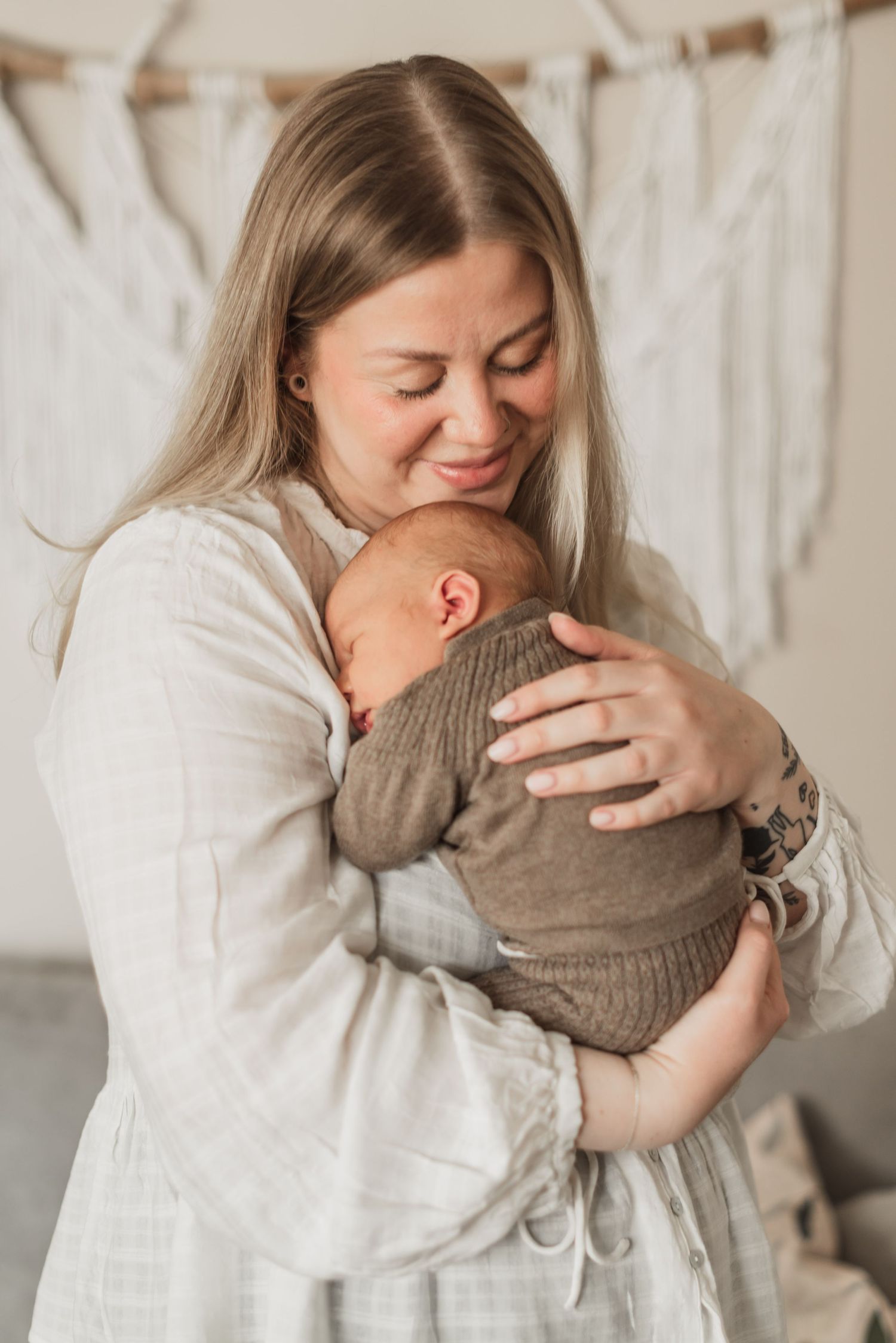 A smiling woman with blonde hair tenderly holds a newborn baby wrapped in a brown knit blanket.