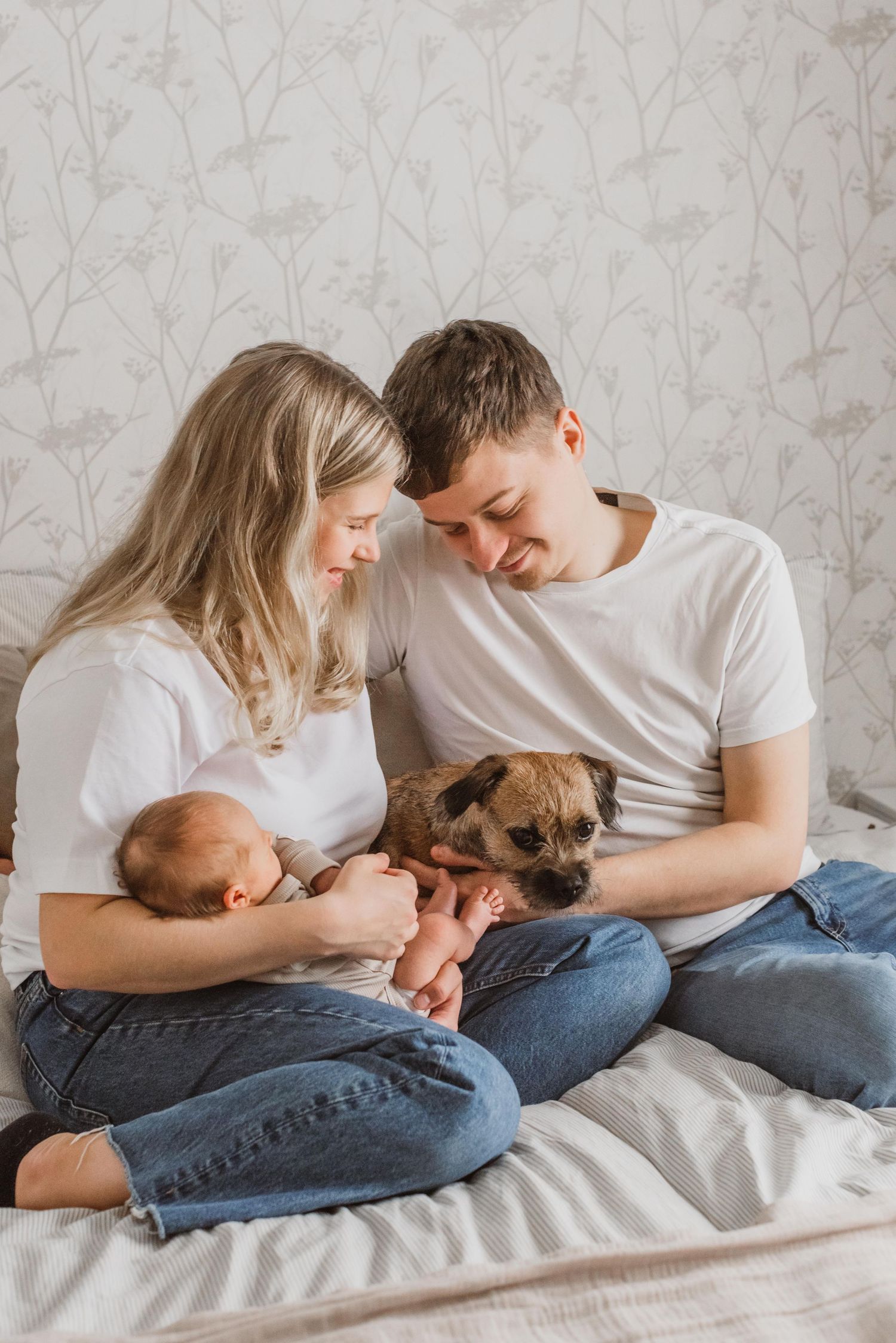 A couple in white shirts with their newborn baby snuggle together with their pug dog on a bed.