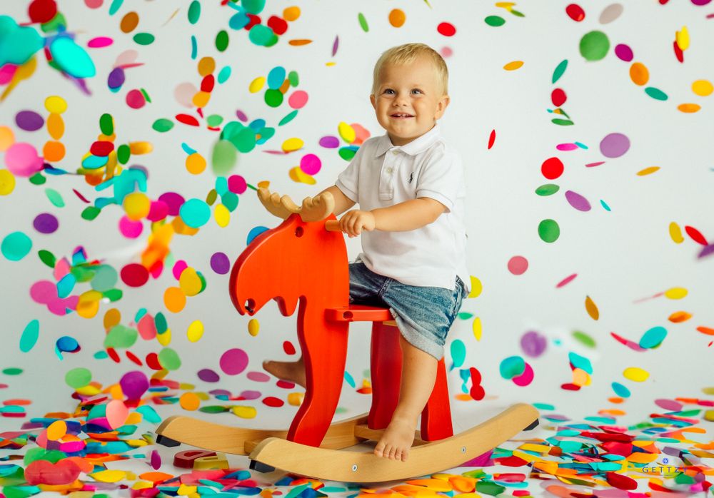 Child rides red rocking horse toy amid colorful confetti shower against white background.