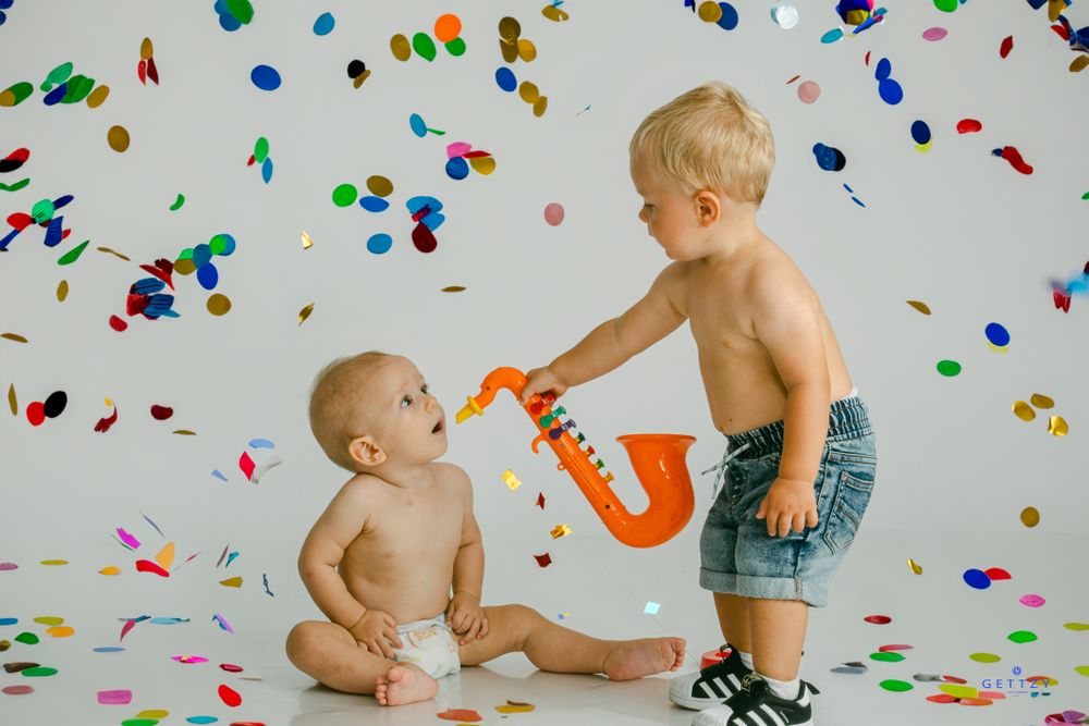 Two young children playing with a toy saxophone amidst colorful confetti on a light gray backdrop during a photoshoot.