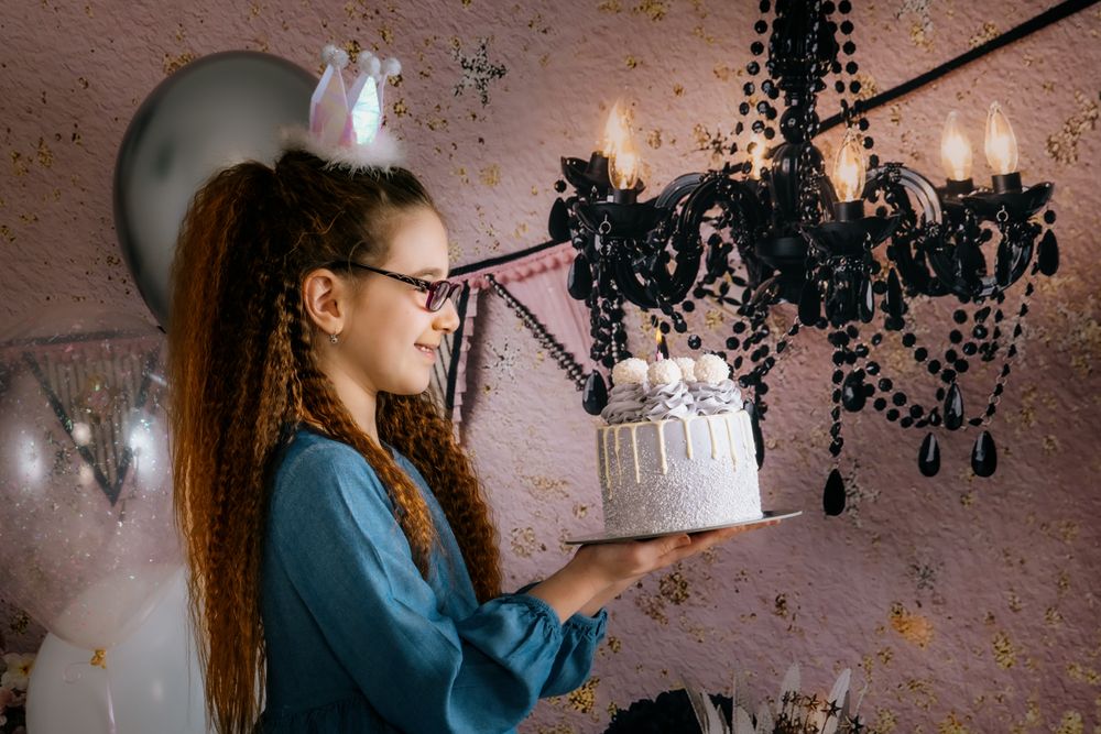 A young person holds a white birthday cake near an elegant black chandelier against a pink textured wall.
