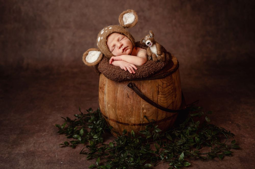 Sleeping baby wearing mouse ears rests peacefully in rustic wooden barrel surrounded by greenery in studio portrait.