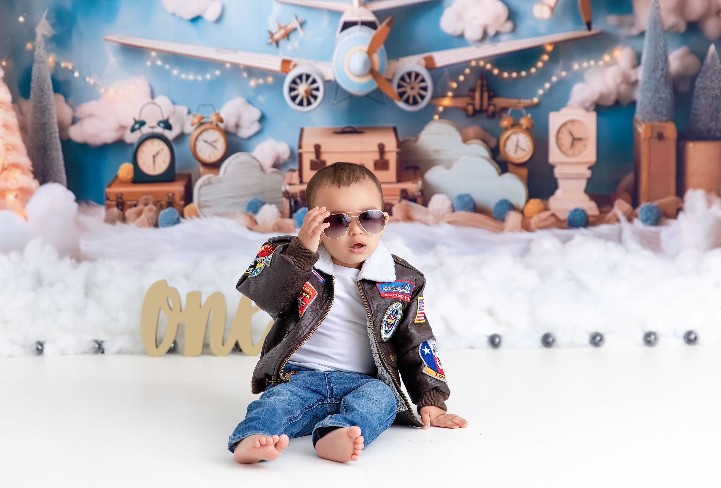 A young child in sunglasses and leather jacket sits in a winter wonderland themed studio setup with clocks and clouds.