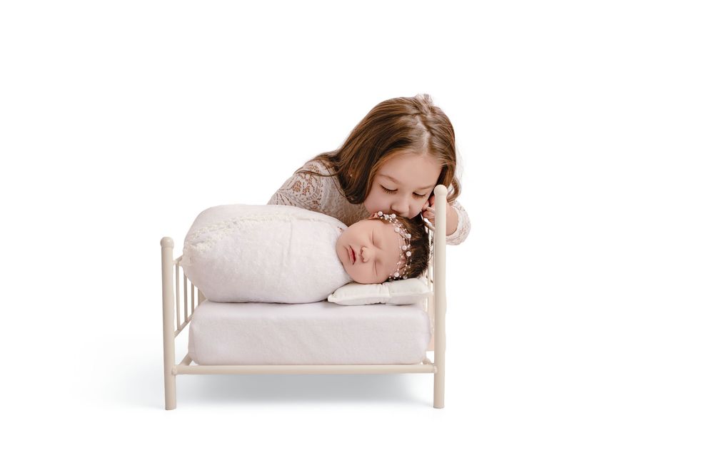 Older sister lovingly kisses sleeping swaddled newborn baby on a white miniature bed in studio.