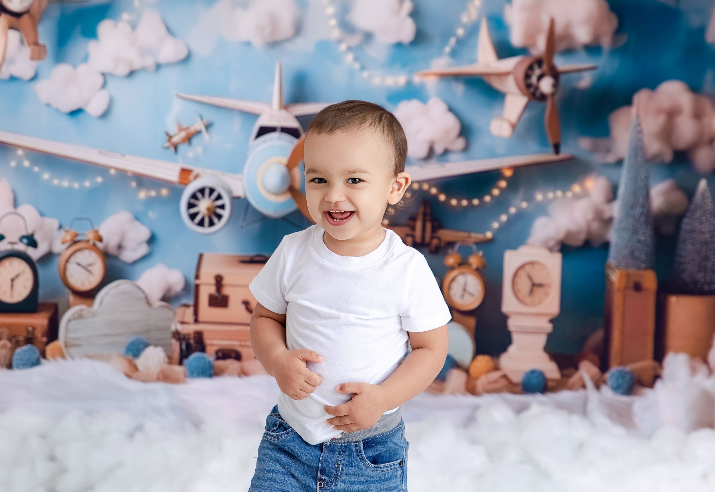 Child smiles in front of whimsical aviation themed backdrop with wooden toy airplanes and fluffy clouds.