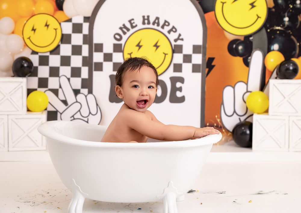 Happy baby sitting in white bathtub during one happy dude birthday cake smash photo session with smiley face balloons.