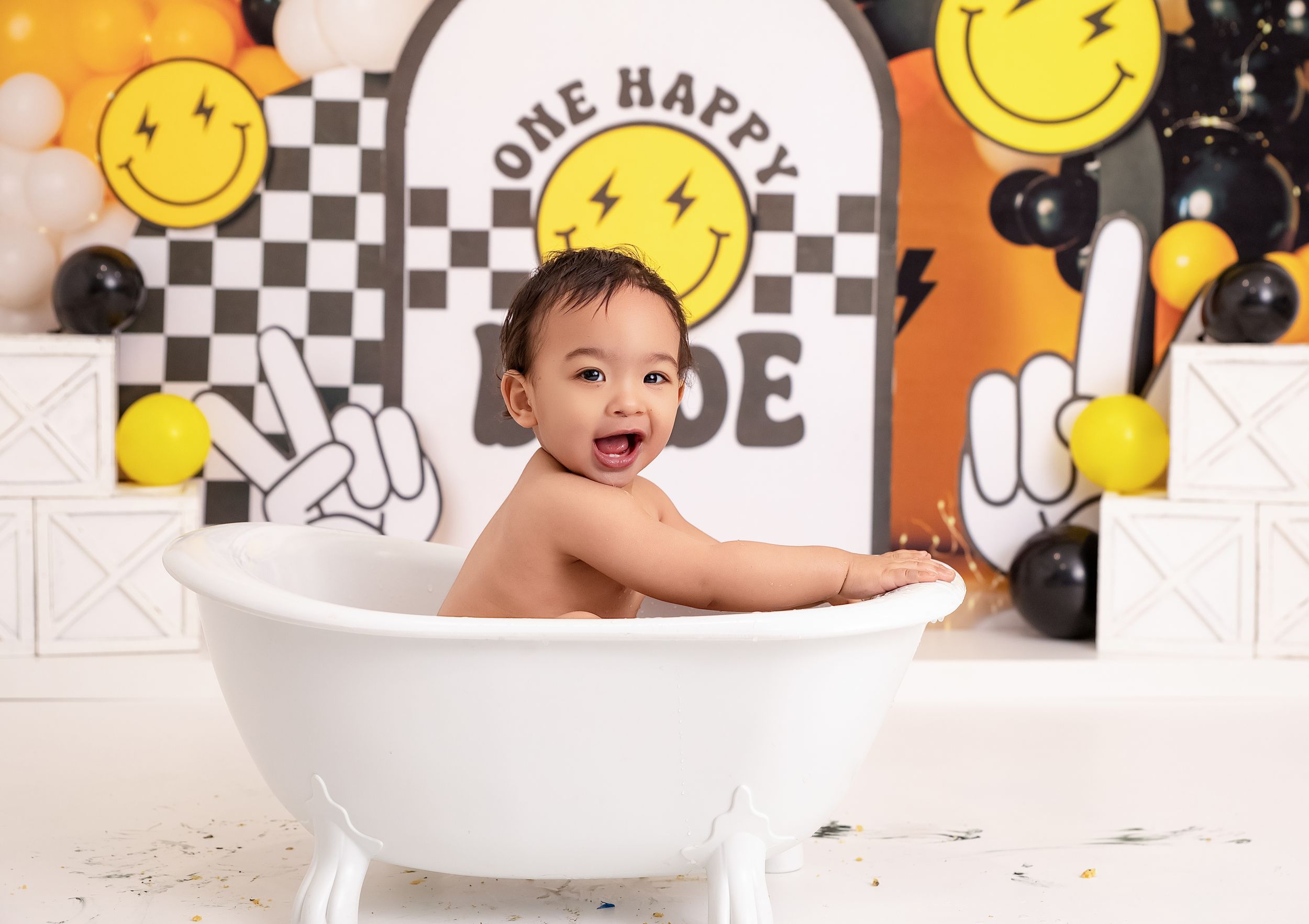 Happy baby sitting in white bathtub during one happy dude birthday cake smash photo session with smiley face balloons.
