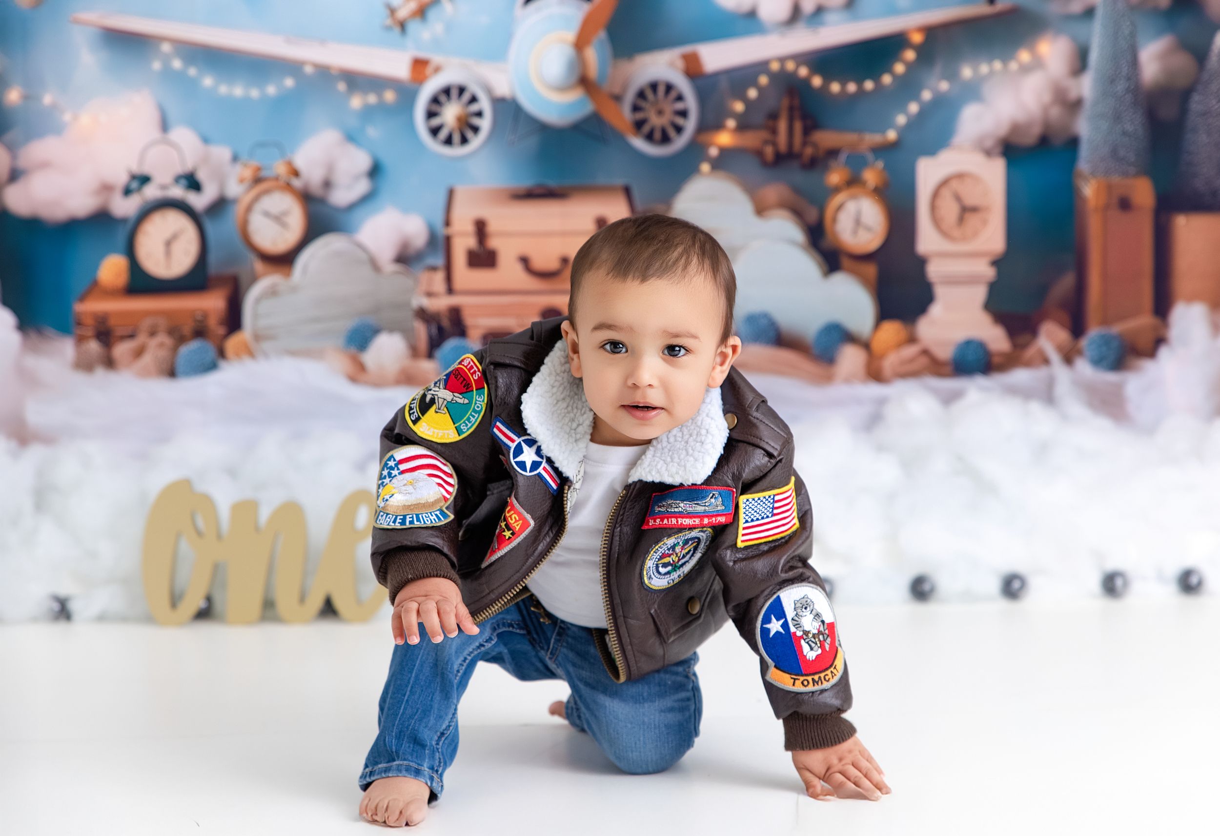 Small child crawls on snowy floor setup with aviation props and decorative string lights against blue backdrop.