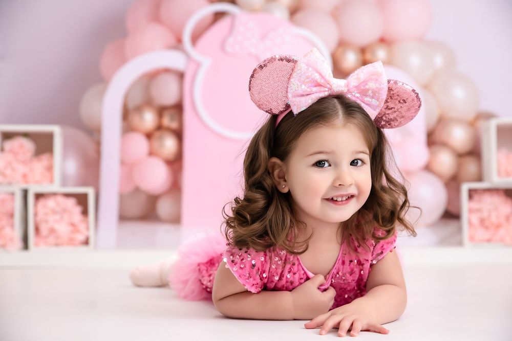 Smiling toddler girl in pink tutu and Minnie Mouse ears poses at a pink balloon birthday party backdrop.