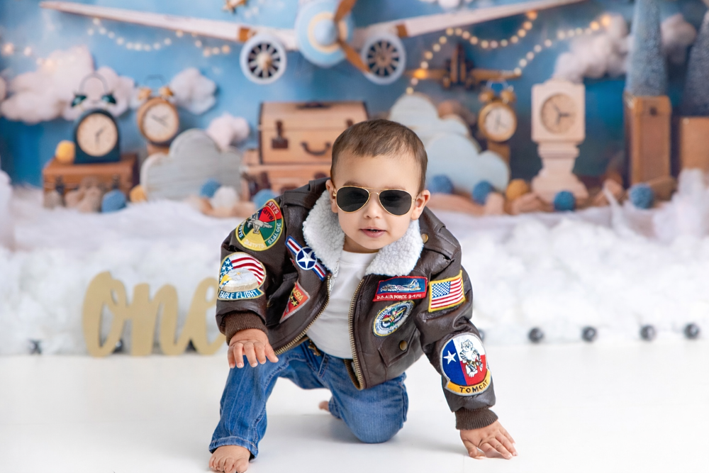 Baby boy in aviator jacket and sunglasses crawling on floor with travel-themed backdrop for first birthday cake smash photo.