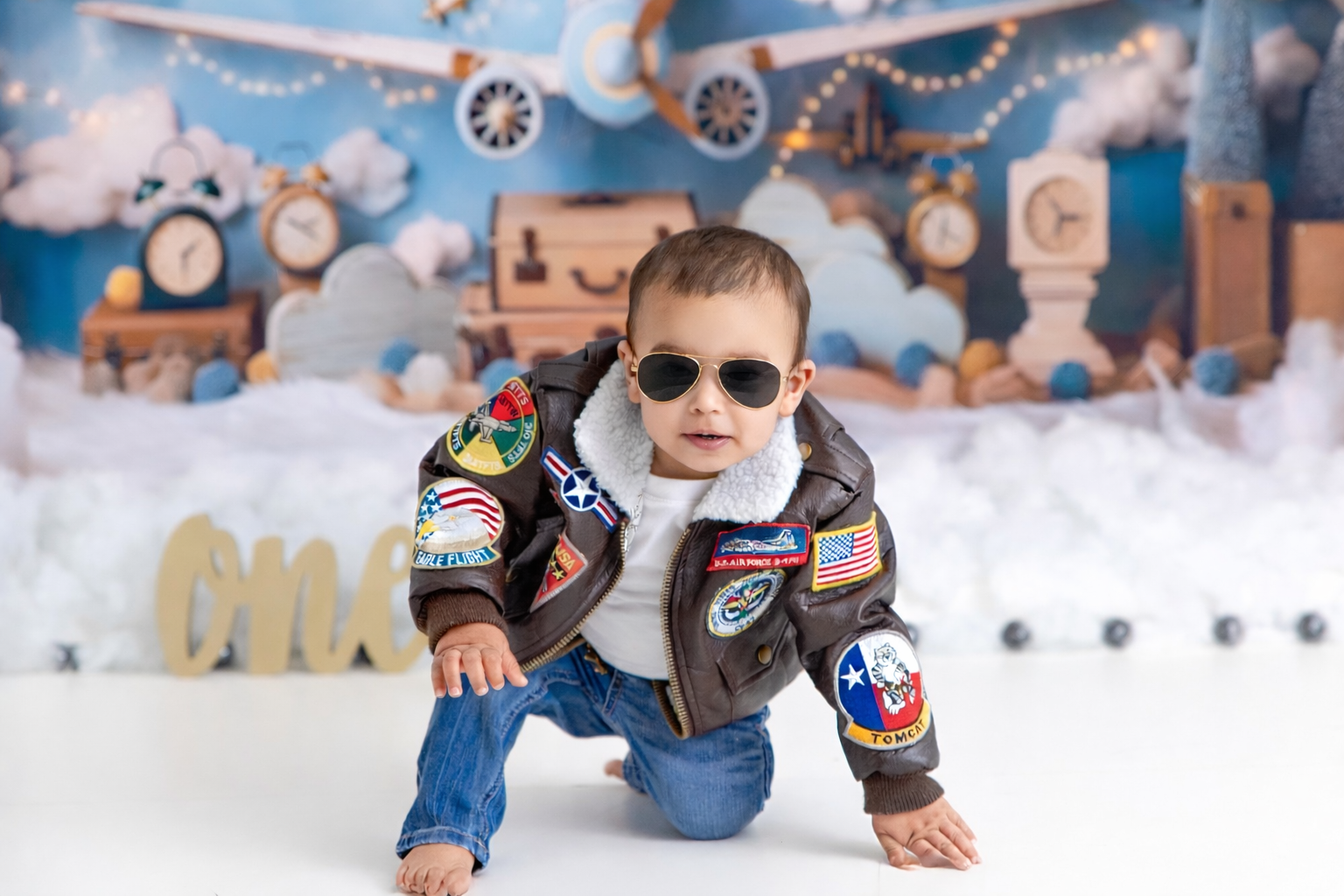 Baby boy in aviator jacket and sunglasses crawling on floor with travel-themed backdrop for first birthday cake smash photo.