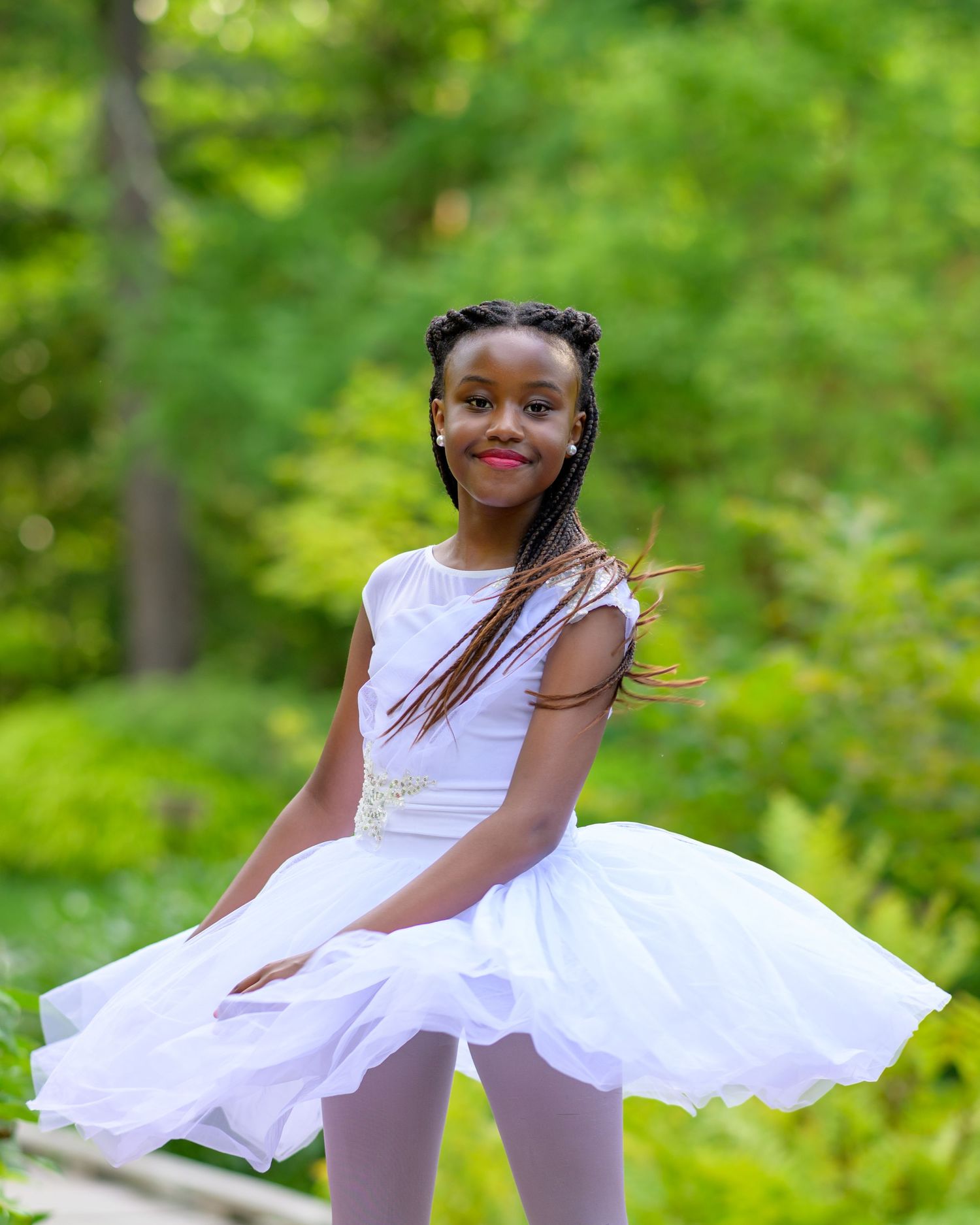 A young African girl with hair braids twirls and poses in her white ballerina tutu on a wooden bridge outdoors in summer.