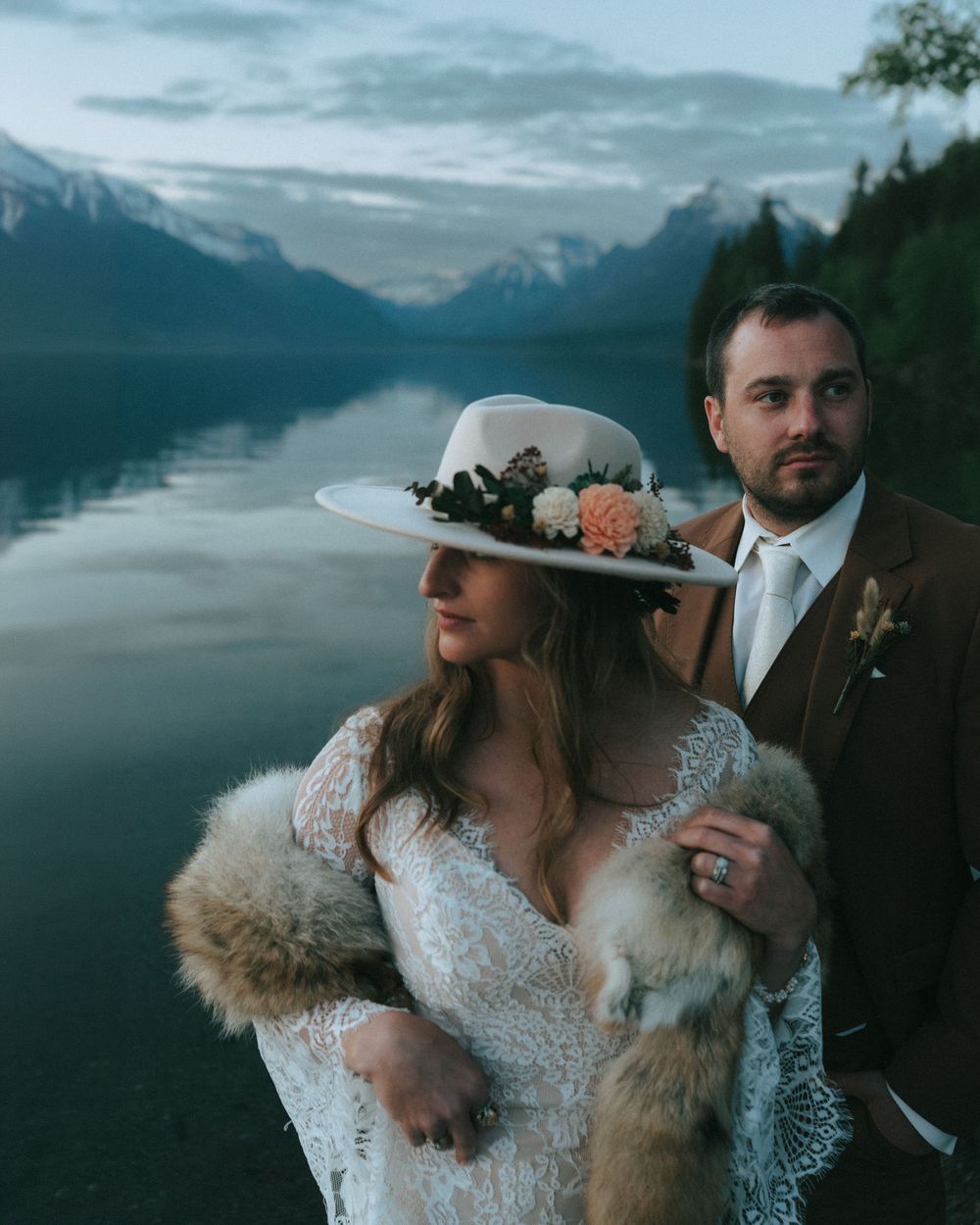 Bride in lace gown, floral hat, fur stole stands with groom in brown suit by mountain lake at dusk.