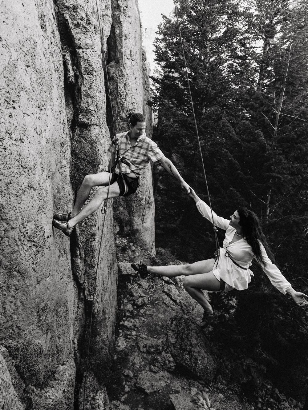 Black and white photo of a couple rock climbing together, holding hands on a steep cliff face surrounded by trees.