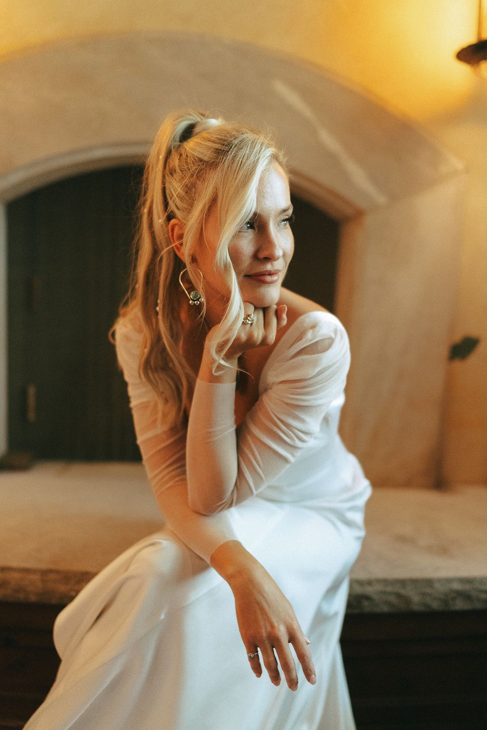 Elegant blonde bride in white dress sitting gracefully, wearing gold earrings in a warmly lit stone interior.