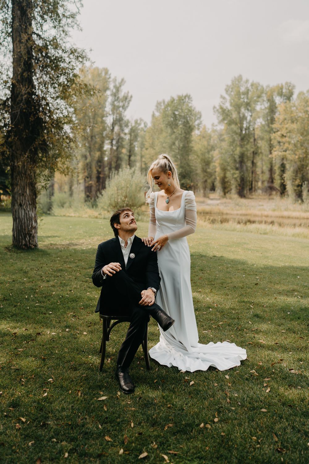Bride in white gown stands beside groom in black suit seated on chair, sharing a loving gaze outdoors.