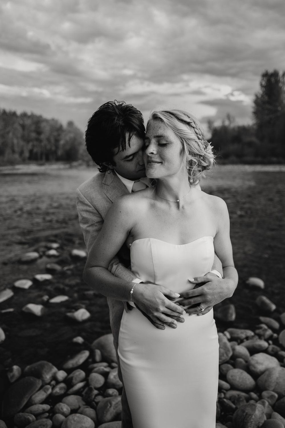 Black and white photo of a groom kissing his bride on the cheek by a rocky riverbank outdoors.