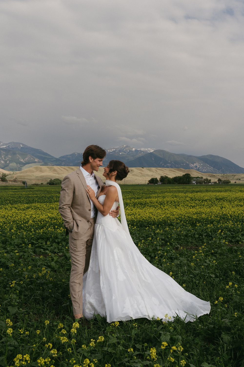 Wedding couple standing in a field of yellow wildflowers with mountain backdrop.