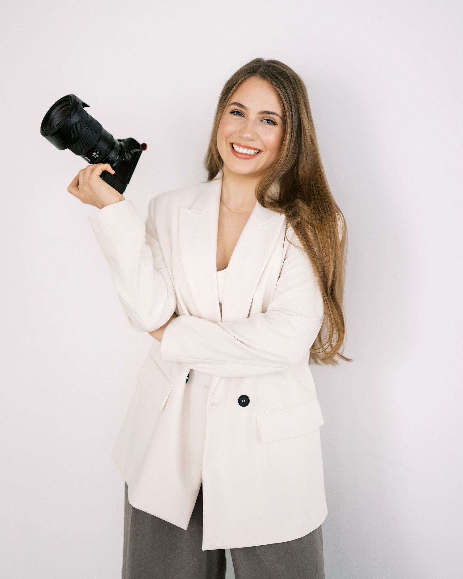 Professional photographer in white blazer smiling while holding a DSLR camera against gray wall.