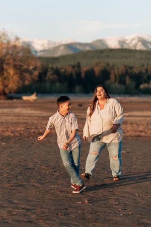 Two people joyfully running together on a beach with snow-capped mountains in the background during golden hour.