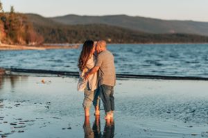 Couple embracing on wet sand at sunset with mountains and water in the background.