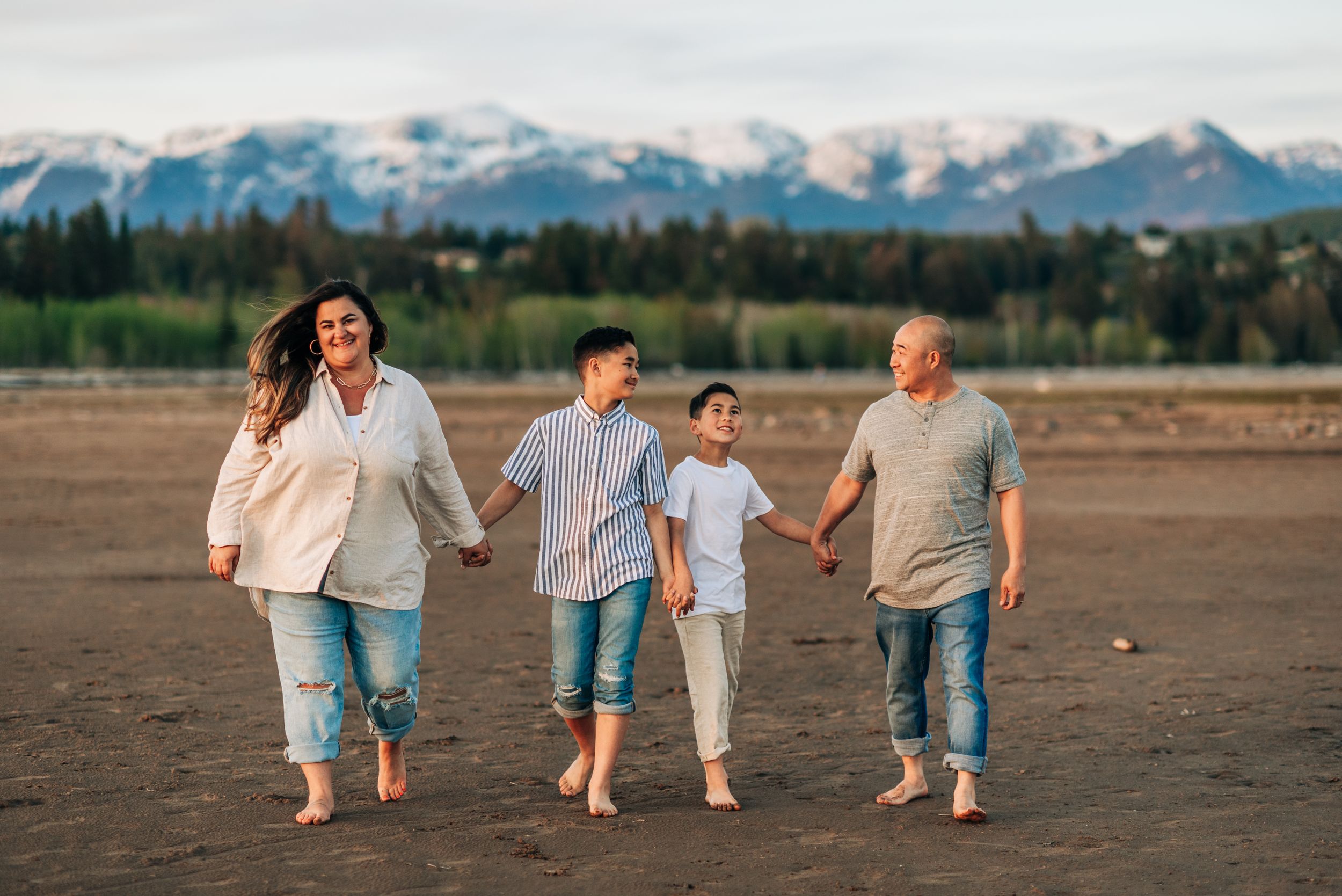 Family of four walking barefoot together on beach with snow-capped mountains in background.
