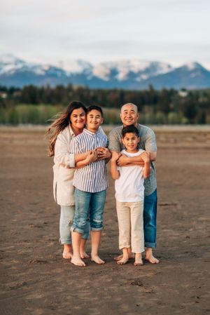 Family standing barefoot together on beach with snow-covered mountains in background.