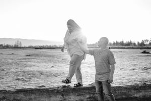 Black and white photo of two people walking on driftwood at the beach.