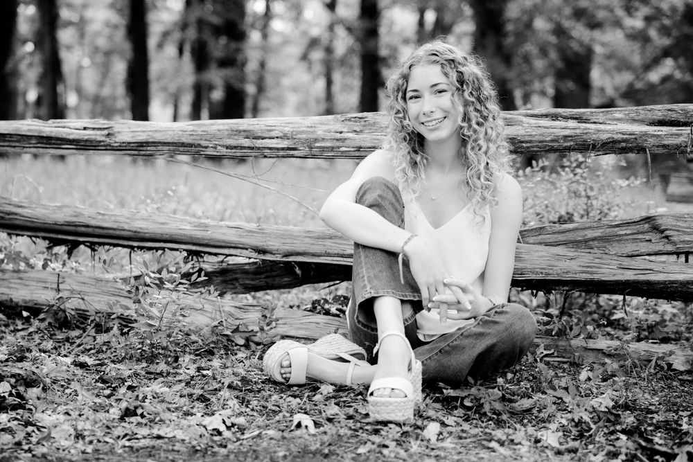 Black and white portrait of someone with curly hair sitting casually on a fallen log in a forest setting.