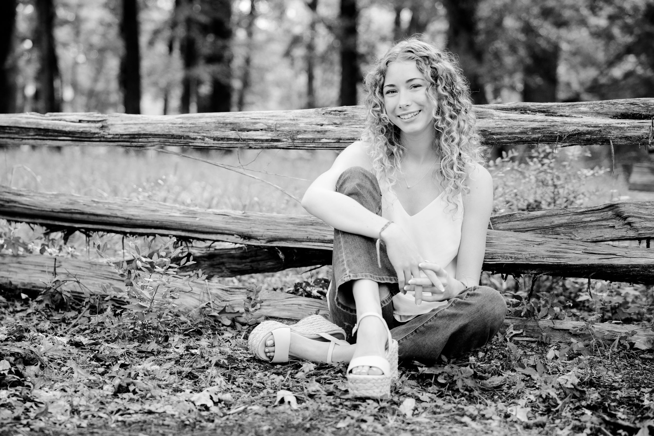 Black and white portrait of someone with curly hair sitting casually on a fallen log in a forest setting.