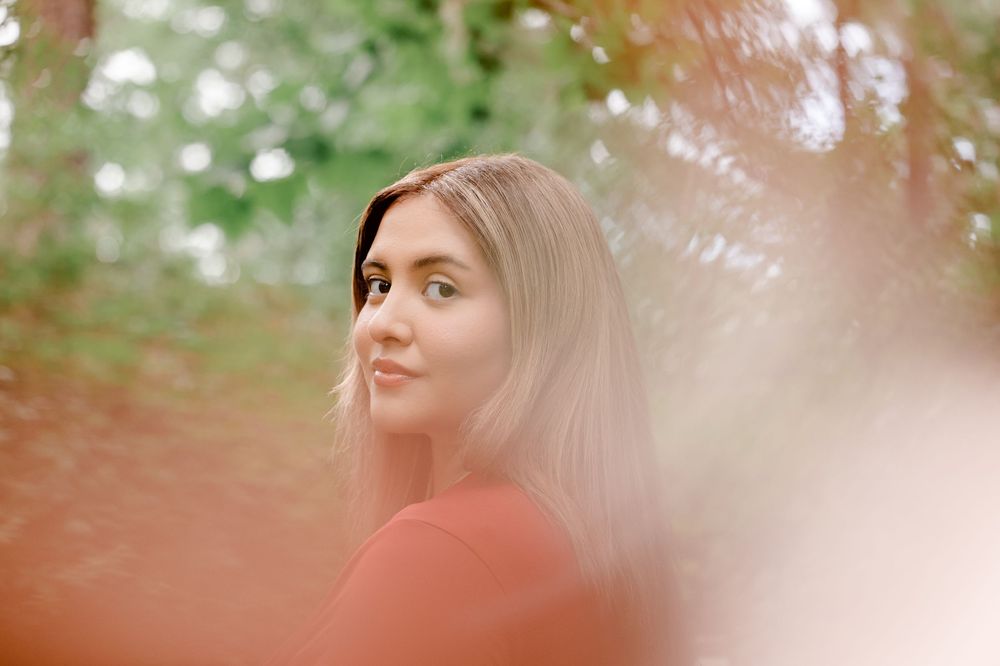 Young woman with blonde hair in a red top, smiling softly among lush green trees in a sunlit forest. Summerville/Charleston, SC