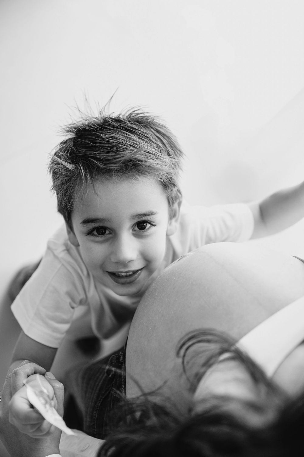 Black and white photo of a smiling young boy leaning on a pregnant woman's belly, holding her hand.