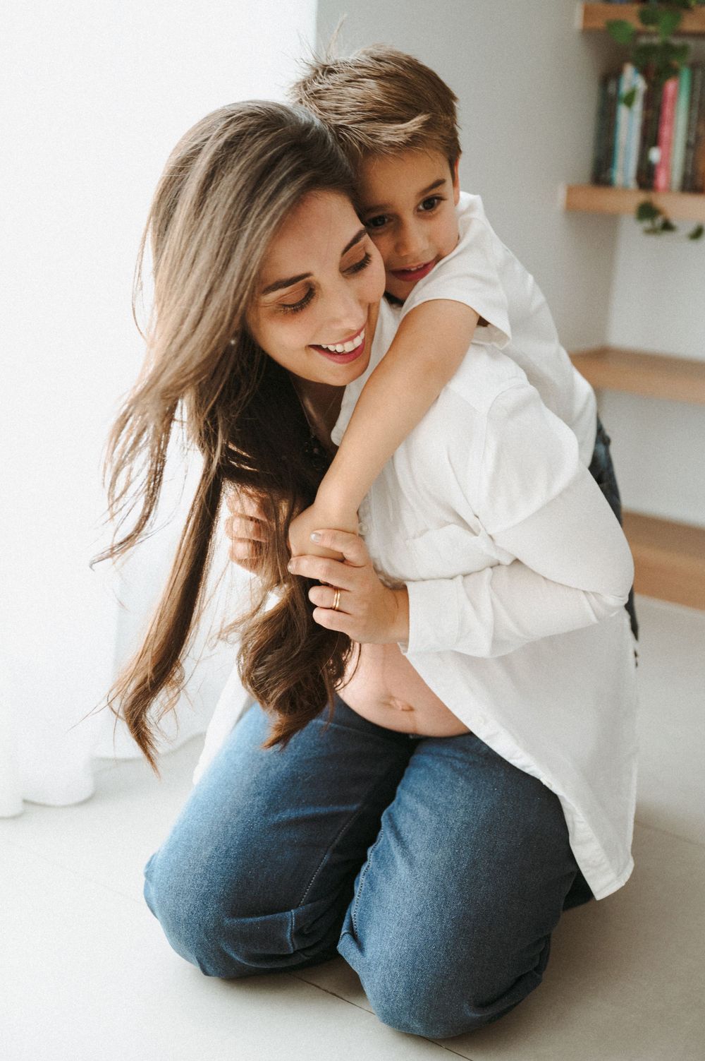 Pregnant woman in white shirt and jeans hugging her young son while sitting on the floor indoors.