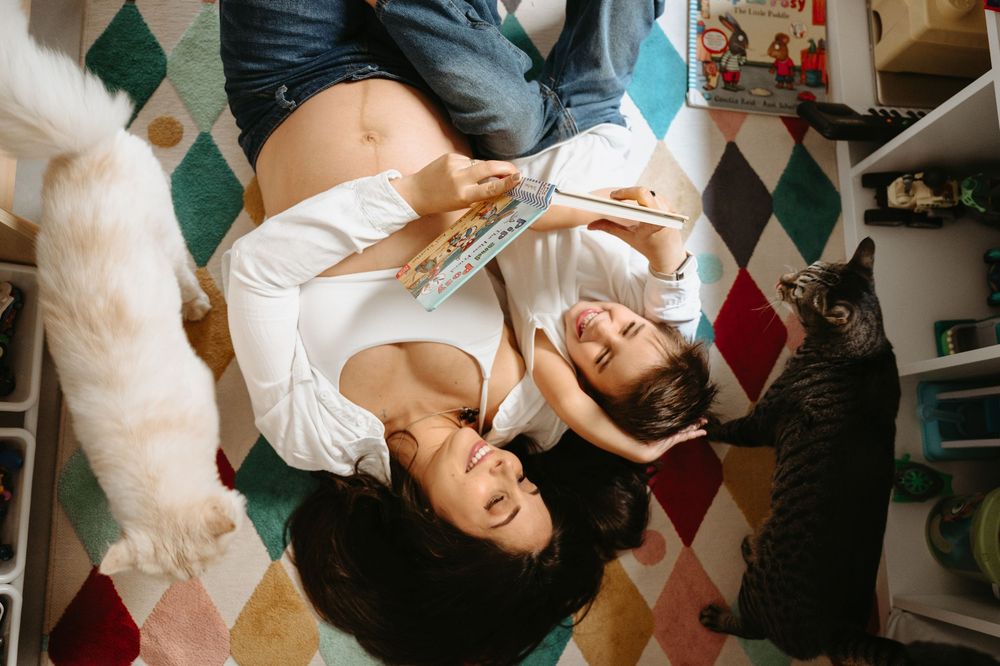 Overhead view of two women lying on a colorful rug, reading a book together, with cats nearby.