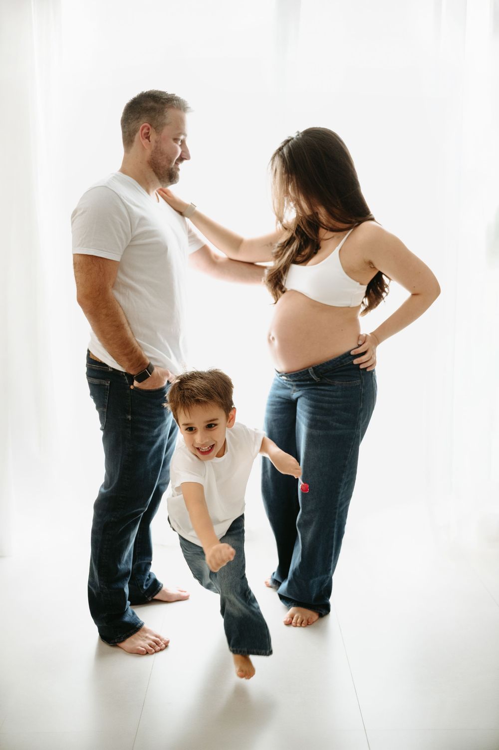 Pregnant woman and man in white shirts and jeans pose with energetic young boy in bright studio setting.