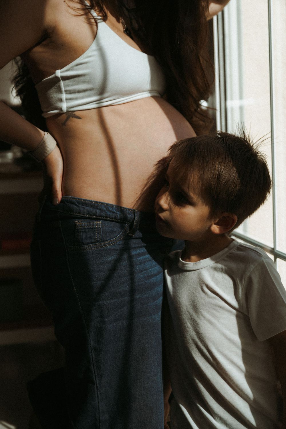 A young child lovingly kisses a pregnant mother's baby bump near a sunlit window.