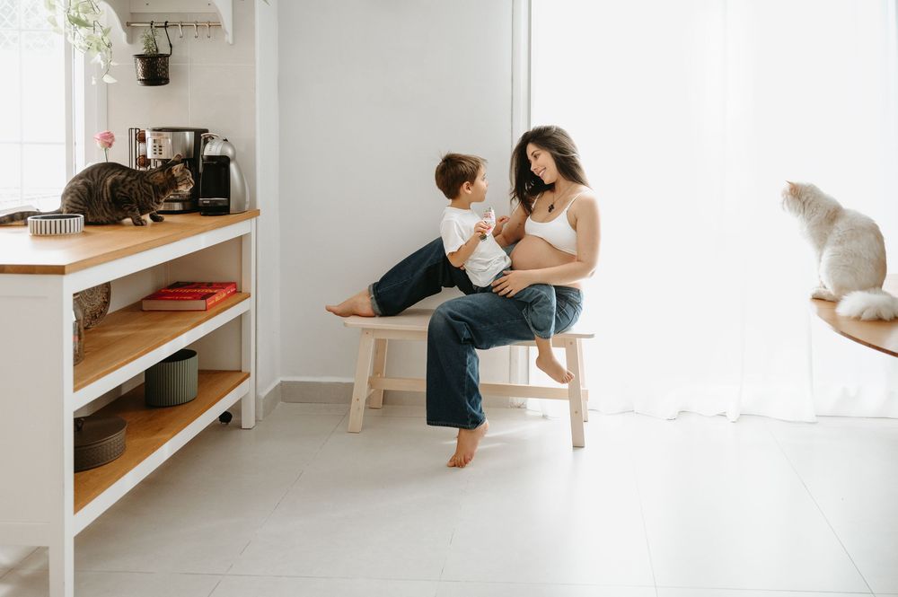 Mother breastfeeding baby on wooden stool in bright kitchen with toddler nearby and cats on shelves.