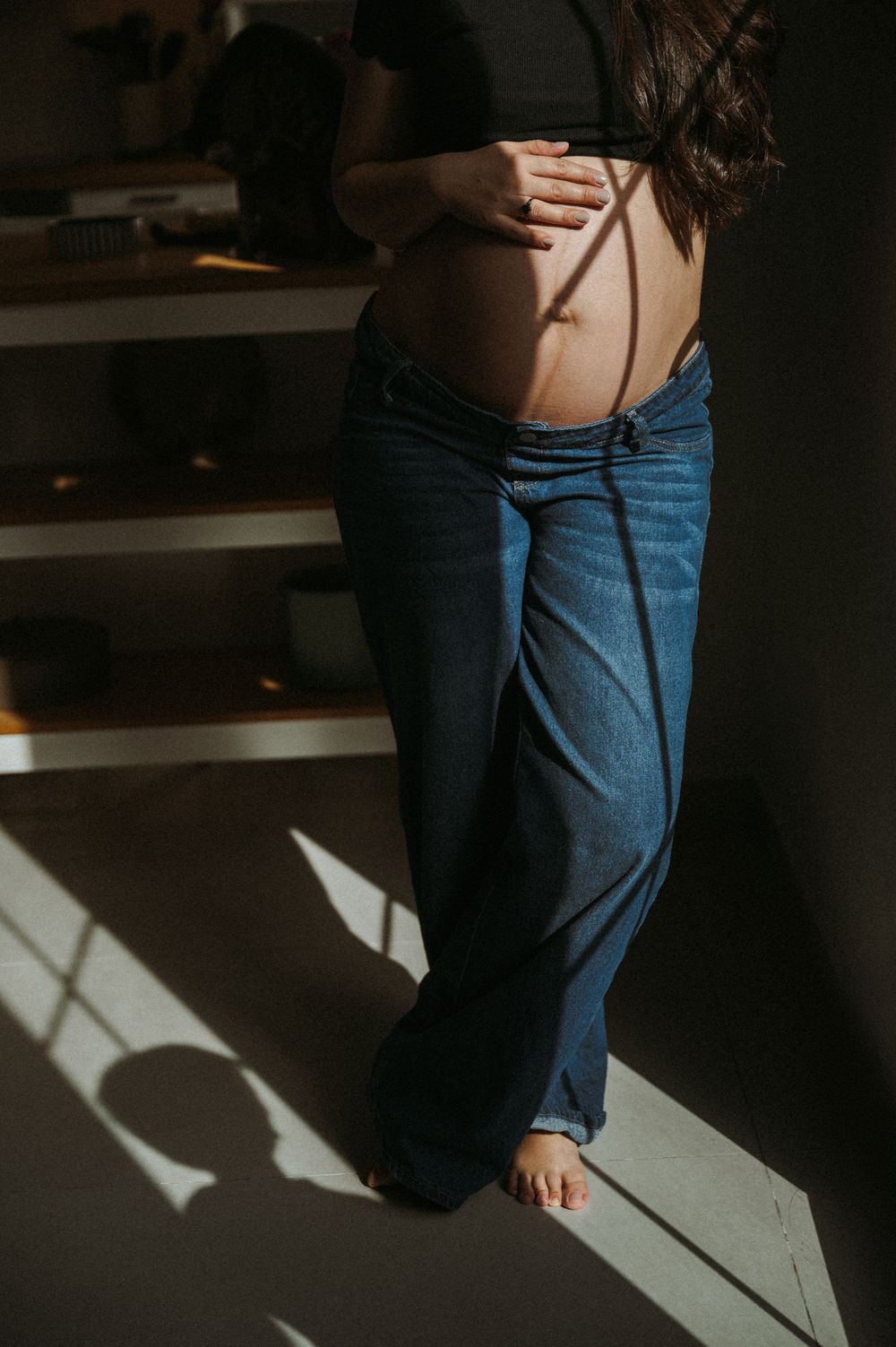 Pregnant woman cradling bare baby bump in loose blue jeans, standing in natural sunlight indoors.